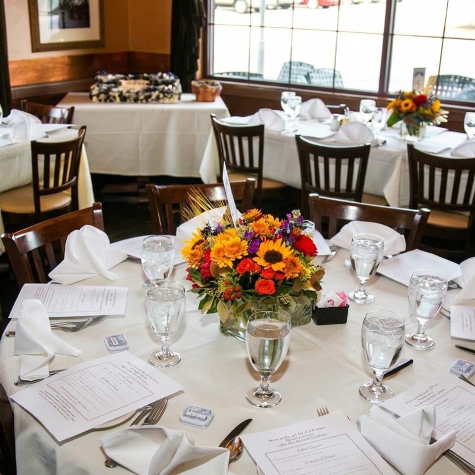 Elegant restaurant dining room with white tablecloths, floral centerpieces, folded napkins, menus, and table settings by large windows.
